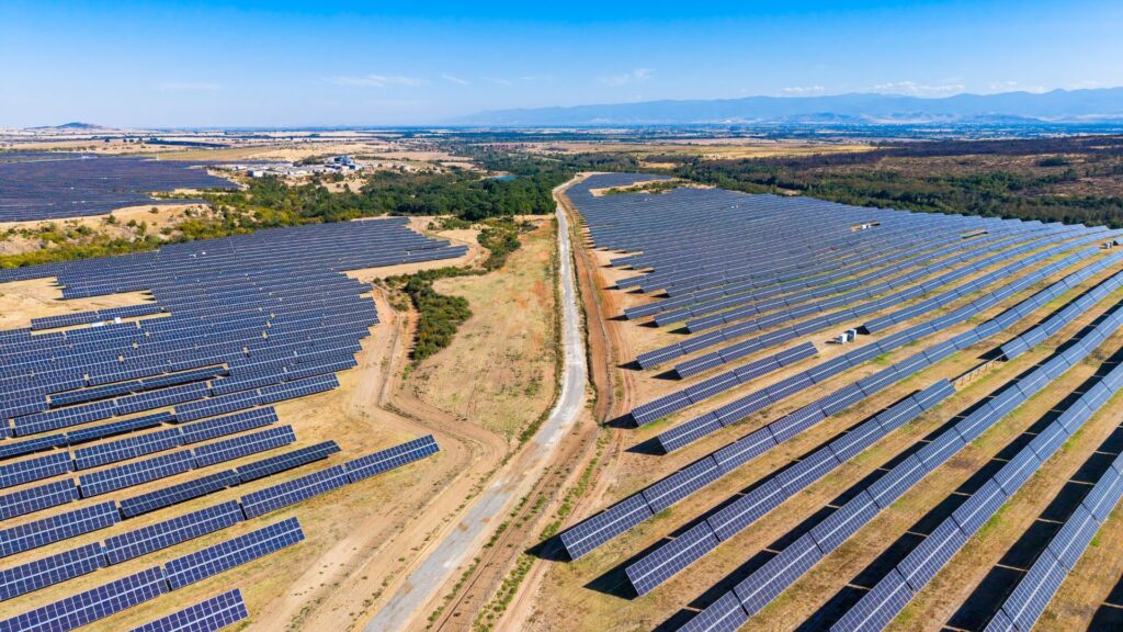 Aerial view of a utility-scale solar farm with rows of photovoltaic panels stretching across a dry landscape, divided by a central access road with mountains in the distance