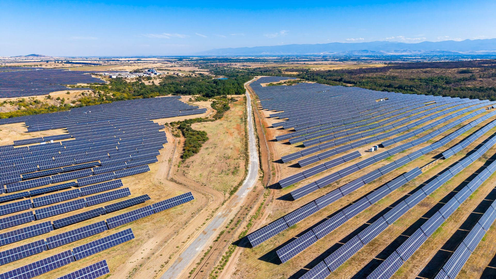 Aerial view of a utility-scale solar farm with rows of photovoltaic panels stretching across a dry landscape, divided by a central access road with mountains in the distance